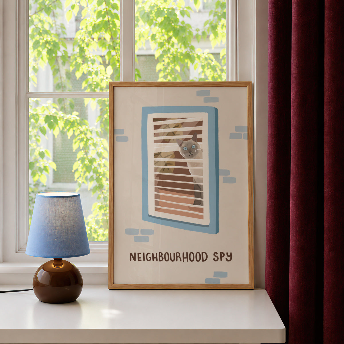 Framed cat spy print with window bars and 'NEIGHBOURHOOD SPY' text, blue accents on tabletop.