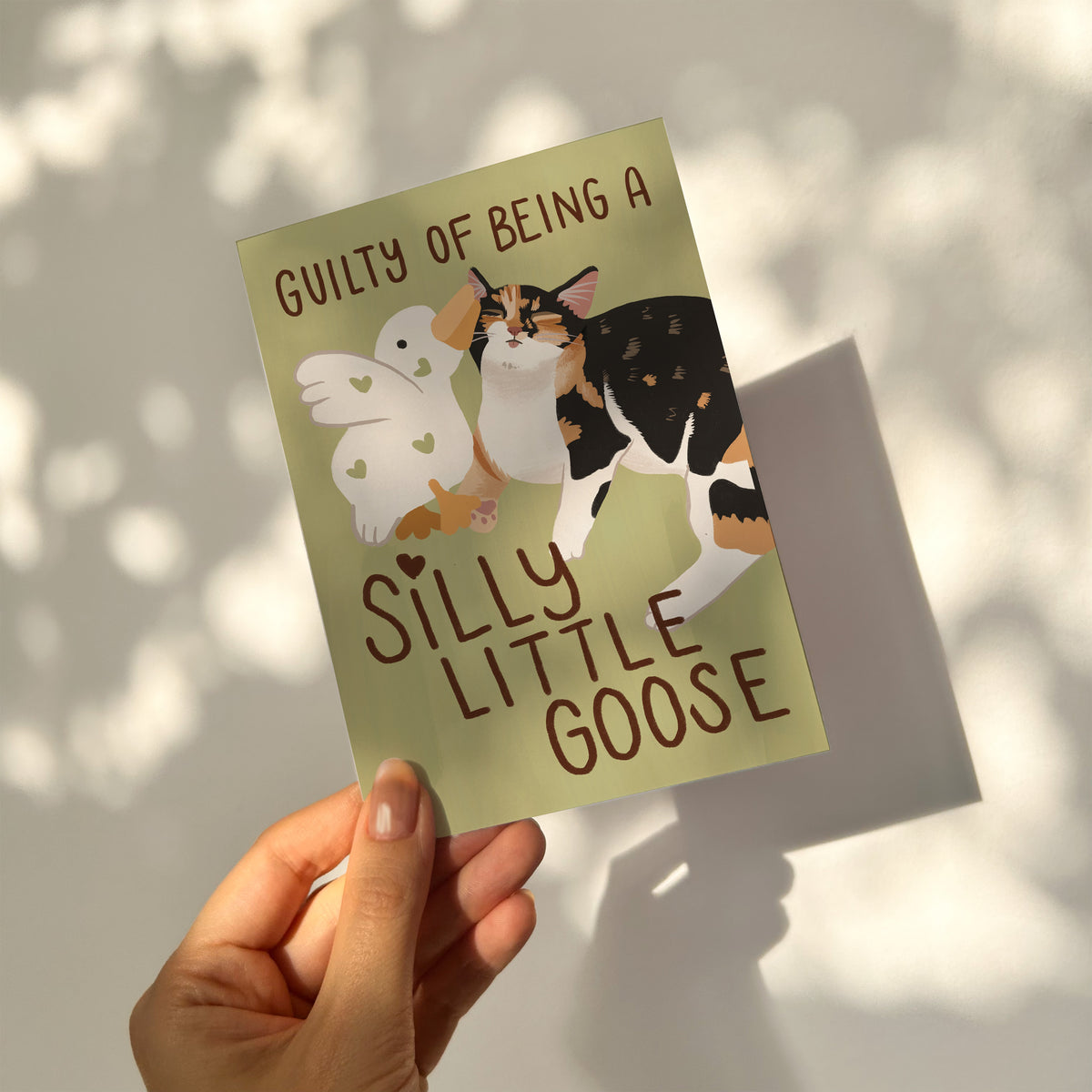Unframed goose and cat illustration print on light green, held up with “Silly Little Goose”
